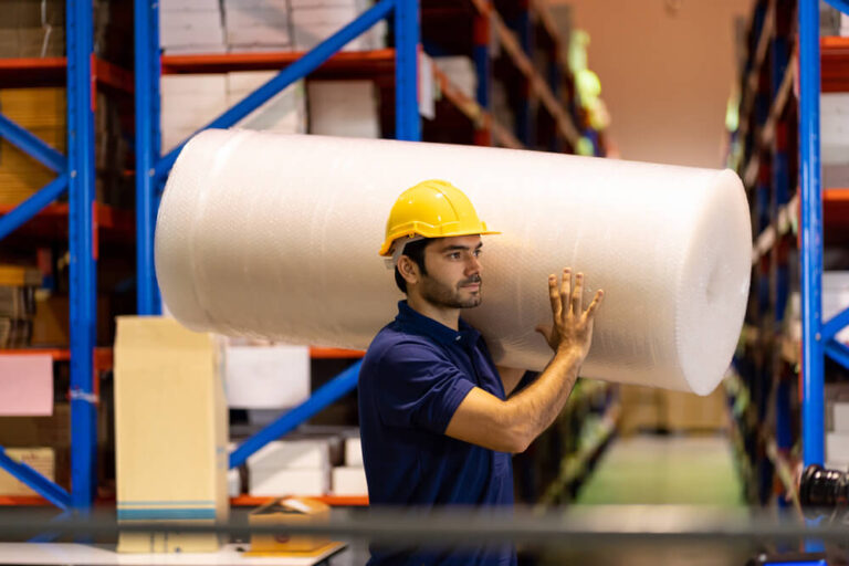 Hombre con uniforme azul y casco amarillo cargando un rollo grande de hule-espuma. Fondo de almacen con racks y cajas Almacén Guadalajara