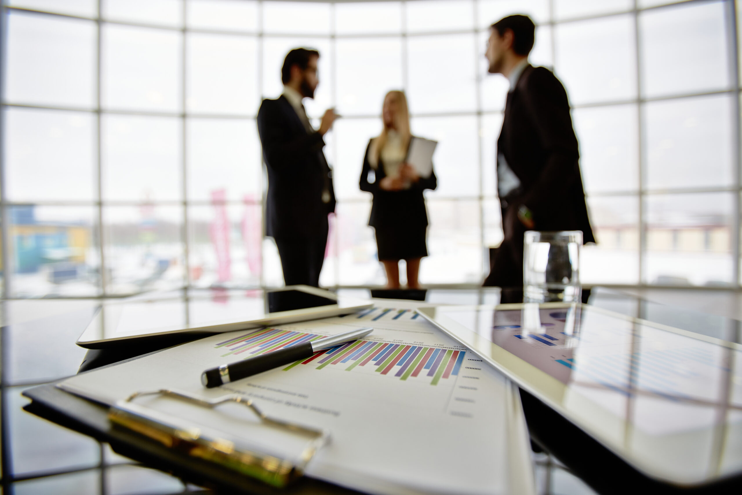 Technological devices and financial document at workplace on background of three business partners discussing ideas